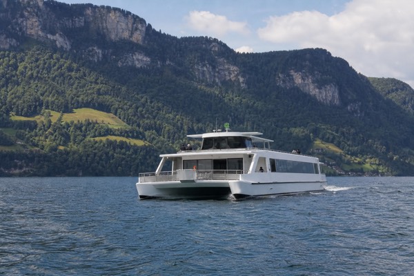 A white twin-hull catamaran crossing Lake Lucerne with a forested ridge behind it