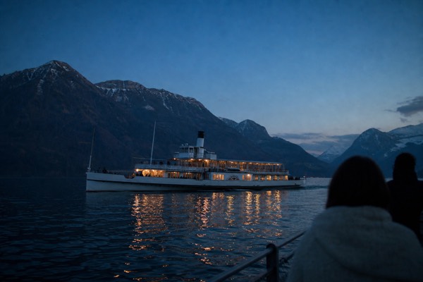A historic paddle steamer with lit windows crossing Lake Lucerne at dusk